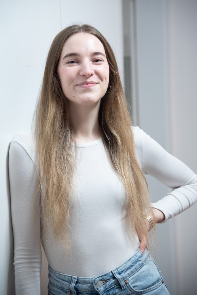 Junge Frau mit langen, glatten Haaren, die in einem weißen Langarmshirt und Jeans steht.