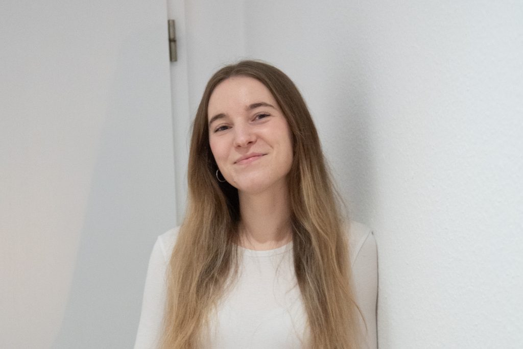 Junge Frau mit langen, glatten Haaren, die an einer weißen Wand steht und lächelt.